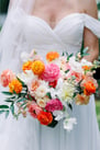 Close-up of a bride in a draped off-the-shoulder white wedding dress, holding a large, vibrant, texturally rich bridal bouquet.