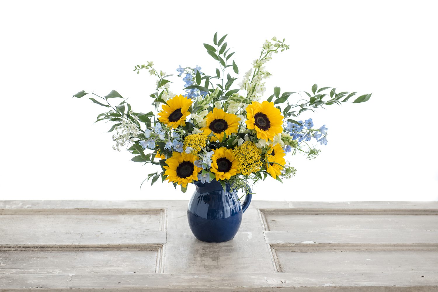 Sunflowers and Delphinium explode out of this blue pitcher vase with greens and Yarrow to complement.