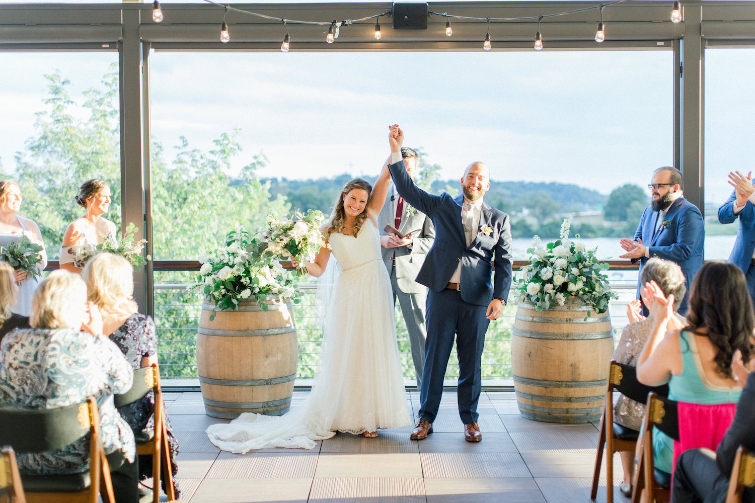 A view of the Anacostia River awaits wedding couples at District Winery in Washington, DC. White and ivory flowers with loose and flirty greens flank the officiant. Image by Abby Grace Photography.
