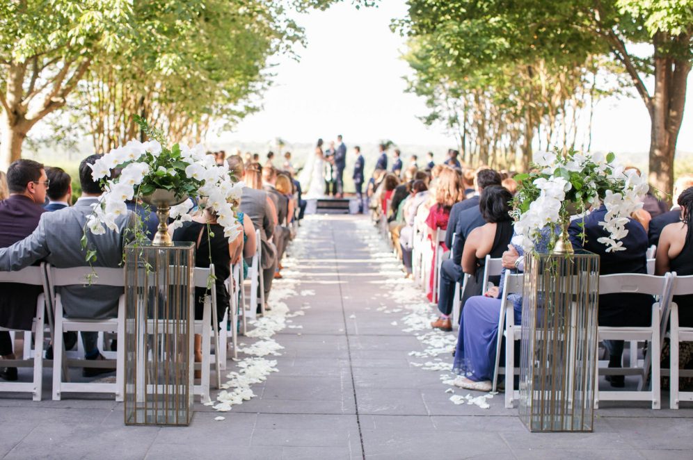 Four arrangements made with Orchids and greenery set on gold and glass stands at the back and front of this ceremony. These were re-purposed on tables set on gold candlestick stands. The petals add a bit of romance at Landsdowne Resort and Spa in Le…