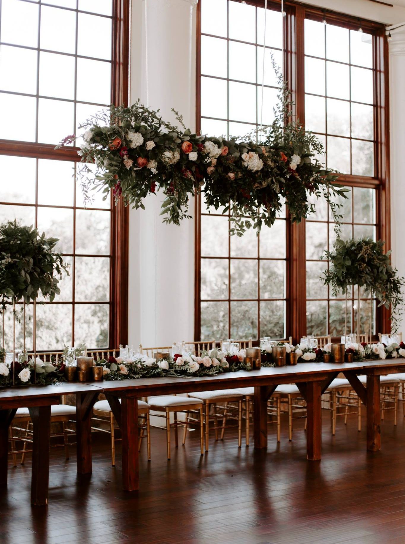 King’s table at Raspberry Plain Manor photographed by We Are the Cashmans. Tall centerpieces with greenery flank the long table with a raised floral design overhead. Floral garland and bridesmaids bouquets with groupings of votive candles.