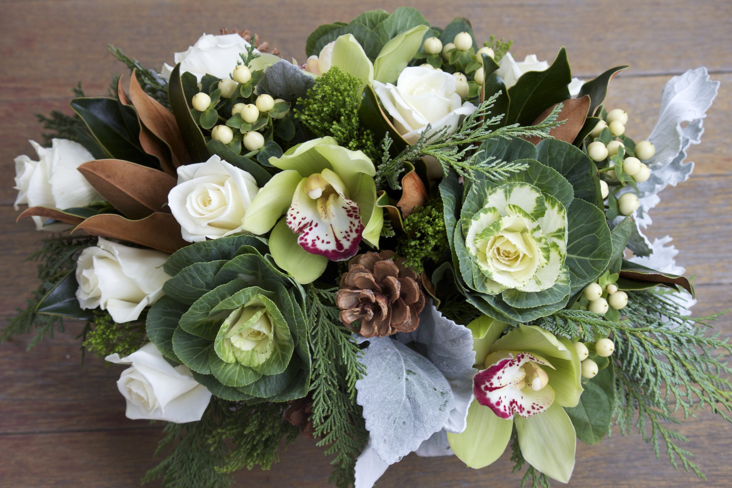 Magnolia leaves (brown), white roses, white Hypericum berries, green Cymbidium orchids (green flower with red tips), decorative cabbage, pine cone, cedar (feather-like greenery), Dusty Miller (gray leaves)